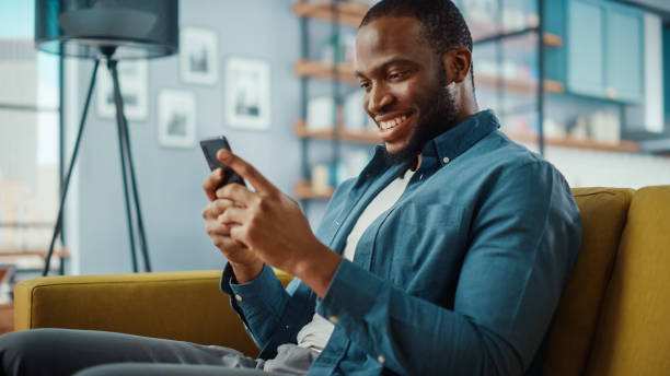 istock Handsome Black African American Man Using Smartphone while Sitting on a Sofa in Cozy Living Room. Freelancer Working From Home. Browsing Internet, Using Social Networks, Having Fun in Flat. 1311087488