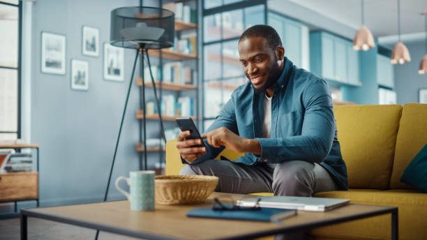 istock Handsome Black African American Man Using Smartphone while Sitting on a Sofa in Cozy Living Room. Freelancer Working From Home. Browsing Internet, Using Social Networks, Having Fun in Flat. 1311088019