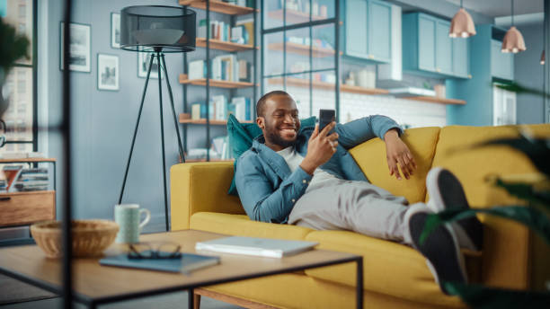 istock Excited Black African American Man Using Smartphone while Resting on a Sofa in Living Room. Happy Man Smiling at Home and Chatting to Colleagues and Clients Over the Internet. Using Social Networks. 1311088023