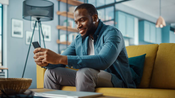 istock Handsome Black African American Man Using Smartphone while Sitting on a Sofa in Cozy Living Room. Freelancer Working From Home. Browsing Internet, Using Social Networks, Having Fun in Flat. 1311088024