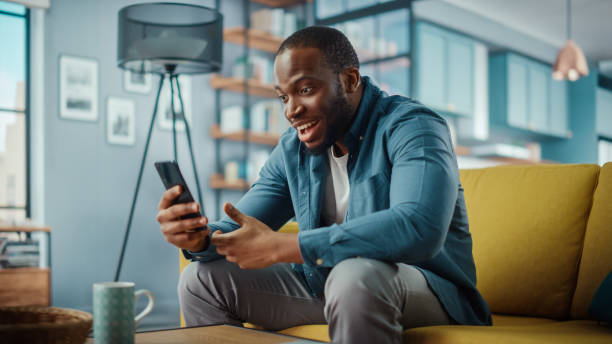 istock Excited Black African American Man Having a Video Call on Smartphone while Sitting on a Sofa in Living Room. Happy Man Smiling at Home and Talking to His Friends and Family Over the Internet. 1311088046