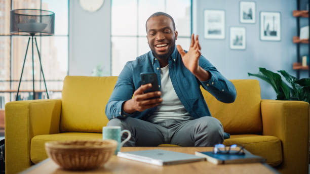 istock Happy Black African American Man Having a Video Call on Smartphone while Sitting on a Sofa in Living Room. Excited Man Smiling at Home and Talking to Colleagues and Clients Over the Internet. 1311088054
