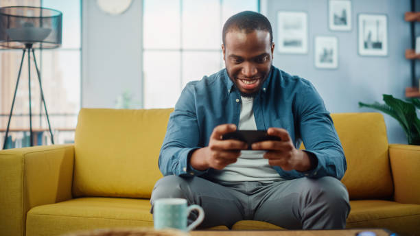 istock Happy Black African American Man Having a Playing Video Game on Smartphone App while Sitting on a Sofa in Living Room. Excited Person of Color Resting at Home and Having Fun Over the Internet. 1311088064