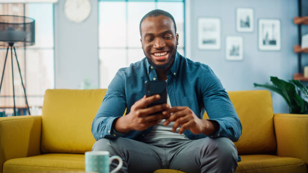 istock Excited Black African American Man Using Smartphone while Resting on a Sofa in Living Room. Happy Man Smiling at Home and Chatting to Colleagues and Clients Over the Internet. Using Social Networks. 1311088082