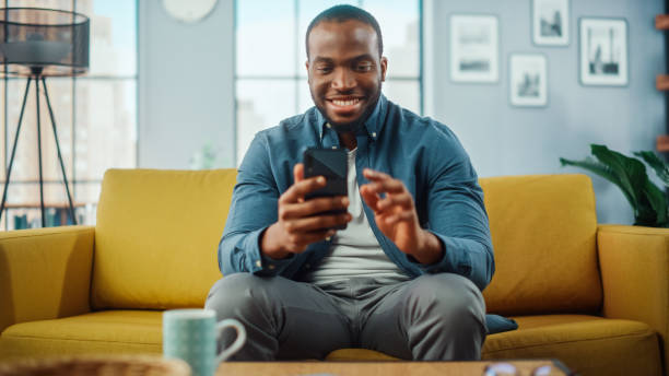 istock Excited Black African American Man Using Smartphone while Resting on a Sofa in Living Room. Happy Man Smiling at Home and Chatting to Colleagues and Clients Over the Internet. Using Social Networks. 1311088083