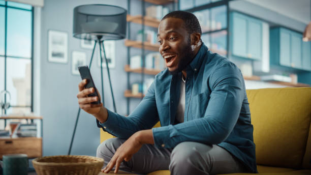 istock Excited Black African American Man Having a Video Call on Smartphone while Sitting on a Sofa in Living Room. Happy Man Smiling at Home and Talking to His Friends and Family Over the Internet. 1311088091
