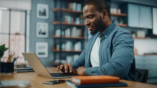 handsome black african american man working on laptop computer while sitting behind desk in cozy living room. freelancer arbeitet von zu hause aus. surfen im internet, verwenden von sozialen netzwerken, spaß in wohnung. - arbeiten von zuhause stock-fotos und bilder