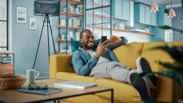 istock Excited Black African American Man Using Smartphone while Resting on a Sofa in Living Room. Happy Man Smiling at Home and Chatting to Colleagues and Clients Over the Internet. Using Social Networks. 1311088111
