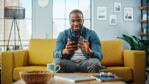 istock Excited Black African American Man Using Smartphone while Resting on a Sofa in Living Room. Happy Man Smiling at Home and Chatting to Colleagues and Clients Over the Internet. Using Social Networks. 1311088117