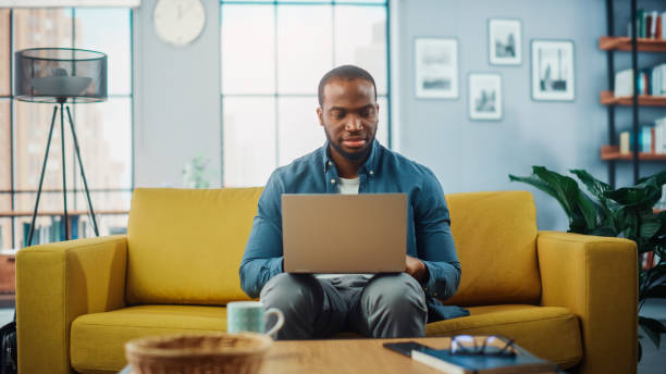istock Handsome Black African American Man Working on Laptop Computer while Sitting on a Sofa in Cozy Living Room. Freelancer Working From Home. Browsing Internet, Using Social Networks, Having Fun in Flat. 1311088122