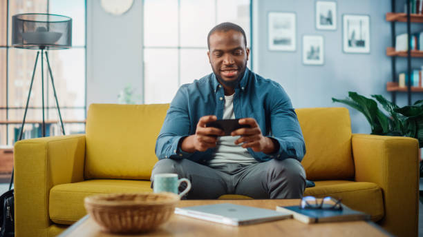 istock Happy Black African American Man Having a Playing Video Game on Smartphone App while Sitting on a Sofa in Living Room. Excited Person of Color Resting at Home and Having Fun Over the Internet. 1311088139