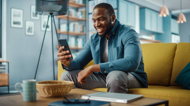 거실에서 소파에 앉아있는 동안 스마트 폰에 비디오 통화를하는 흥분 흑인 아프리카 계 미국인 남자. 집에서 미소짓고 인터넷을 통해 친구와 가족에게 이야기하는 행복한 남자. - 휴대전화 뉴스 사진 이미지