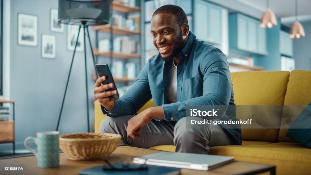 興奮的非洲裔黑人男子坐在客廳的沙發上， 在智慧手機上進行視頻通話。快樂的人在家裡微笑，通過互聯網與朋友和家人交談。 - 免版稅人圖庫照片