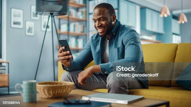 興奮的非洲裔黑人男子坐在客廳的沙發上 在智慧手機上進行視頻通話快樂的人在家裡微笑通過互聯網與朋友和家人交談 照片檔及更多 人 照片