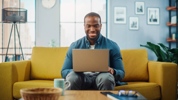 istock Handsome Black African American Man Working on Laptop Computer while Sitting on a Sofa in Cozy Living Room. Freelancer Working From Home. Browsing Internet, Using Social Networks, Having Fun in Flat. 1311088148