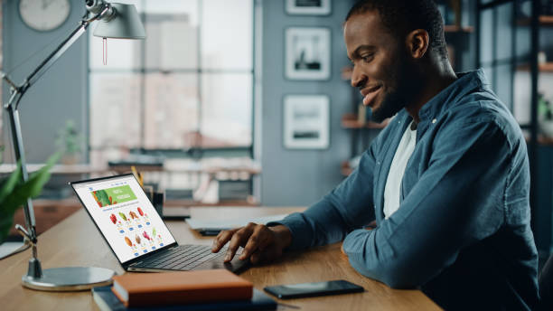 Handsome Black African American is Using Laptop Computer with Groceries Delivery Web Page to Order Fresh Vegetables to Make Dinner. He Lives in Modern Huge Appatment.
