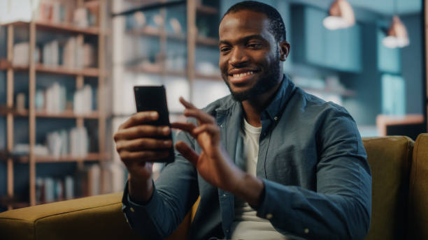istock Excited Black African American Man Using Smartphone while Resting on a Sofa in Living Room. Happy Man Smiling at Home and Chatting to Colleagues and Clients Over the Internet. Using Social Networks. 1311088190