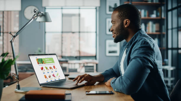 istock Handsome Black African American is Using Laptop Computer with Groceries Delivery Web Page to Order Fresh Vegetables to Make Dinner. He Lives in Modern Huge Appatment. 1311088201