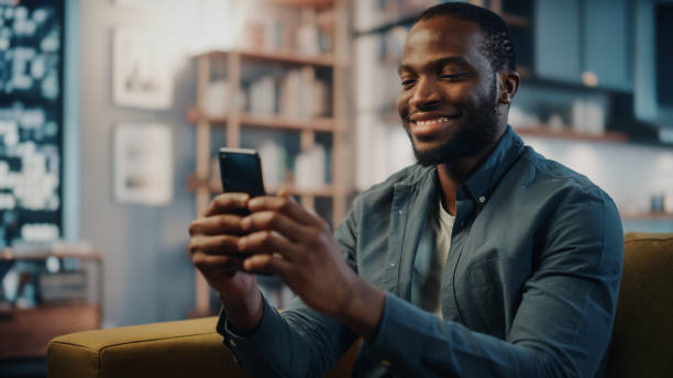 istock Excited Black African American Man Using Smartphone while Resting on a Sofa in Living Room. Happy Man Smiling at Home and Chatting to Colleagues and Clients Over the Internet. Using Social Networks. 1311088209