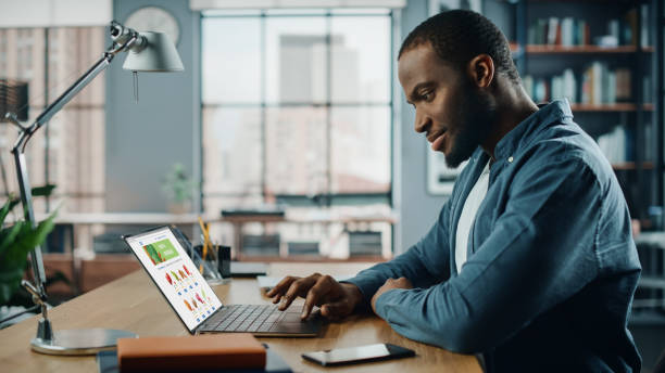 istock Handsome Black African American is Using Laptop Computer with Groceries Delivery Web Page to Order Fresh Vegetables to Make Dinner. He Lives in Modern Huge Appatment. 1311088211