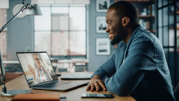 istock Handsome African American Man Having a Video Call with Female Doctor on Laptop Computer while Sitting Behind Desk in Living Room. Freelancer Working From Home and Talking a Medic Over the Internet. 1311088230