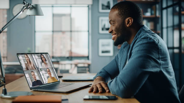 handsome african american man having a video call on laptop computer while sitting behind desk in living room. freelancer working from home and talking to colleagues and clients over the internet. - colleague horizontal business construction imagens e fotografias de stock