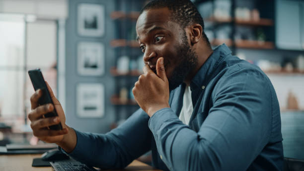 istock Shocked Black African American Man Using Smartphone while Sitting at Table in Living Room. Man Recived Terrable News while Chatting to Family Members and Friends Over the Internet or Social Networks. 1311088296