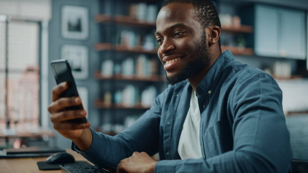 istock Happy Black African American Man Using Smartphone while Sitting at Table in Living Room. Excited Man Smiling at Home and Talking to Colleagues, Clients and Friends Over the Internet. 1311088315