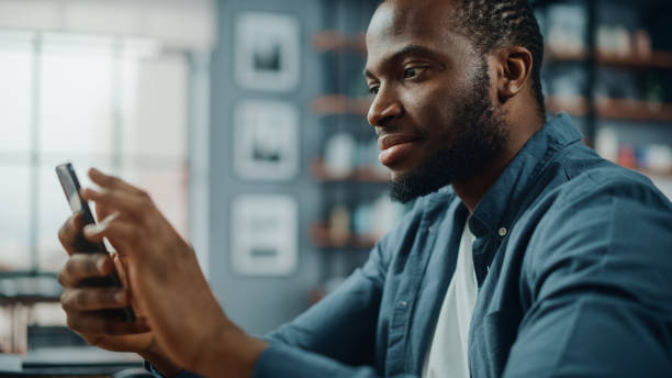 istock Excited Black African American Man Using Smartphone while Sitting at Table in Living Room. Happy Man Smiling at Home and Chatting to Colleagues and Clients Over the Internet. Using Social Networks. 1311088318