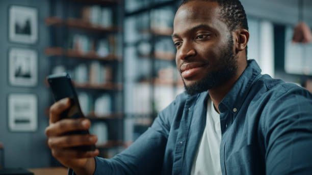 istock Happy Black African American Man Using Smartphone while Sitting at Table in Living Room. Excited Man Smiling at Home and Talking to Colleagues, Clients and Friends Over the Internet. 1311088319