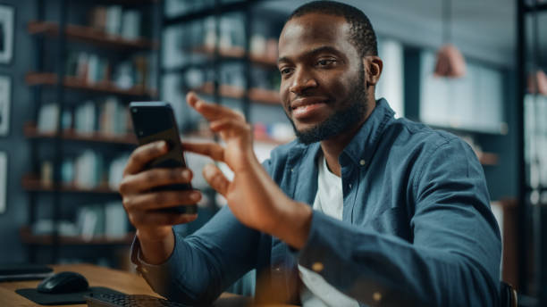 istock Excited Black African American Man Using Smartphone while Sitting at Table in Living Room. Happy Man Smiling at Home and Chatting to Colleagues and Clients Over the Internet. Using Social Networks. 1311088336