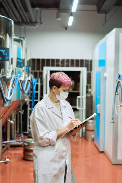 istock Young female brewery expert in whitecoat and protective mask making notes in front of new equipment 1311088970