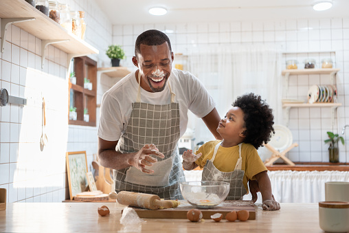 Cheerful smiling Black son enjoying playing with his father while doing bakery at home. Playful African family having fun cooking baking cake or cookies in kitchen together. Single Dad Lifestyle