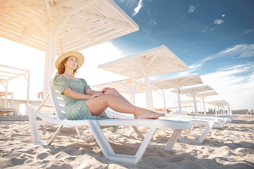 Young attractive caucasian woman in a green dress and a straw sits resting on a lounger under a wooden umbrella on a sandy beach of the seashore or ocean. Rest on the islands of the ocean. Vacation.