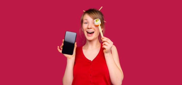 banner,long format. a blonde posing with a smartphone and holding rolls with chopsticks for chinese cuisine. red background with side blank space. - food dinner prepared fish gourmet imagens e fotografias de stock