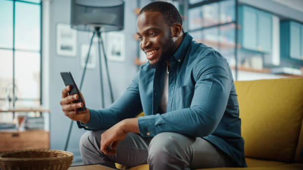 istock Excited Black African American Man Having a Video Call on Smartphone while Sitting on a Sofa in Living Room. Happy Man Smiling at Home and Talking to Colleagues and Clients Over the Internet. 1311093977
