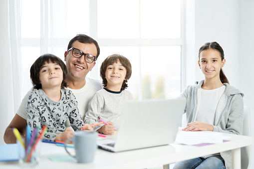 Happy latin father and kids smiling at camera, sitting together at the table at home. Man using laptop, working from home and watching kids. Freelance, family concept