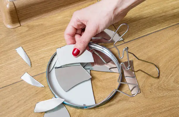 Photo of Woman's hand removes broken pieces of mirror from wooden floor at home