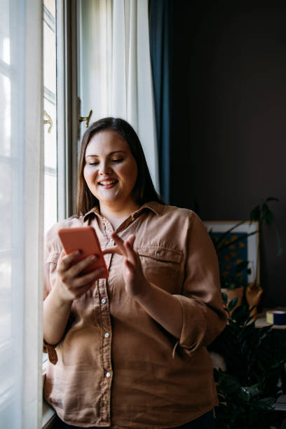istock Beautiful Plus Size Woman Standing next to a Window while Browsing the Internet on her Smartphone 1311096869