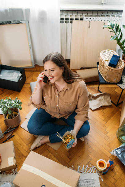 istock Moving in: Happy Plus Size Woman Sitting on the Floor and Talking on her Smartphone while Having a Lunch Break 1311096885