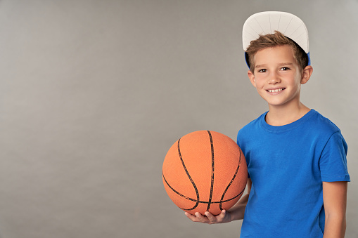 Joyful male child in cap holding basketball ball and smiling while standing against gray background