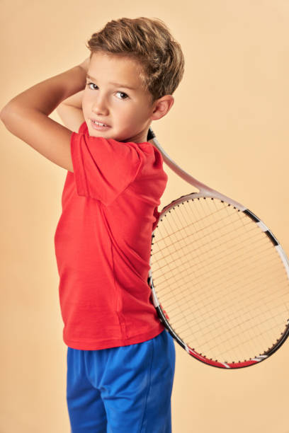 istock Cute boy with tennis racket standing against light orange background 1311097683