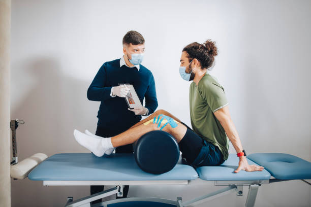 Physical Therapist Holding an Anatomy Chart and Explaining the Problem to the Patient Athlete is sitting on the bed with pillow below his knees listening to the physical therapist explaining knee anatomy to him on the chart and what is a problem with his knee. sports medicine photos stock pictures, royalty-free photos & images