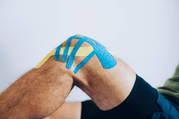 Human Knee with Applied Kinesio Tape Close up of the knee of male athlete with blue and yellow kinesio tape applied to it man sitting with a bandage on his leg stock pictures, royalty-free photos & images