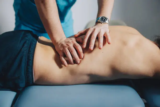 Physical therapist is using his hands to massage the lower back of a male patient lying on his stomach.