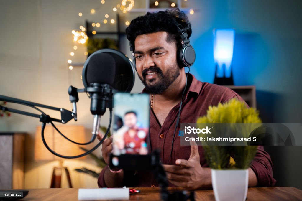 Young social media influencer recording his podcast on mobile phone - concept of vlogging, content creation from home office. Young social media influencer recording his podcast on mobile phone - concept of vlogging, content creation from home office Influencer Stock Photo