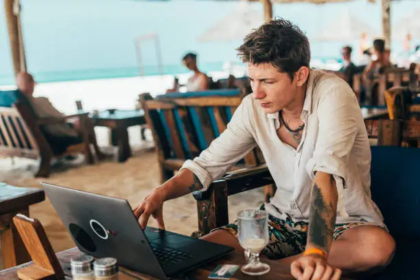 A young handsome man works remotely on a laptop on the internet and drinks coffee in a cafe on the beach.
Working while travelling. Summer vacation in Zanzibar