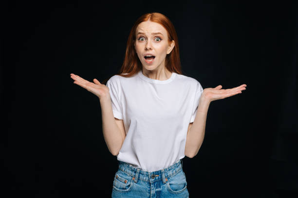 confused young woman wearing t-shirt and denim spreads hands to sides and shrugs shoulders - blank expression head and shoulders horizontal studio shot imagens e fotografias de stock