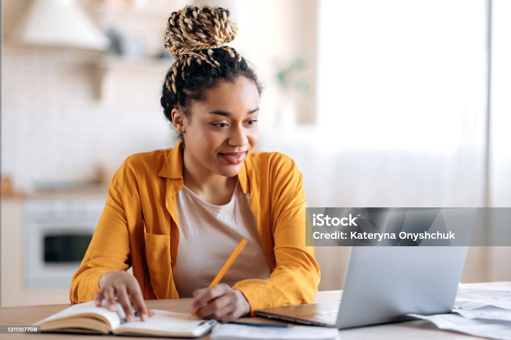 Focused cute stylish african american female student with afro dreadlocks, studying remotely from home, using a laptop, taking notes on notepad during online lesson, e-learning concept, smiling Learning Stock Photo