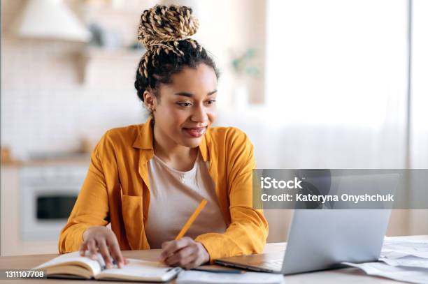 Focused Cute Stylish African American Female Student With Afro Dreadlocks Studying Remotely From Home Using A Laptop Taking Notes On Notepad During Online Lesson Elearning Concept Smiling Stock Photo - Download Image Now